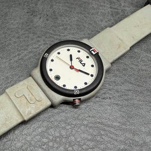 May include: A vintage Fila watch with a light gray band and a black bezel. The watch face is white with black numerals and the Fila logo. The watch has red and black hands and a red button.