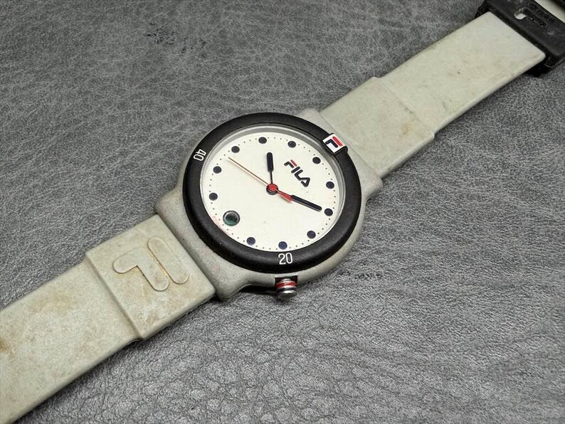 May include: A vintage Fila watch with a light gray band and a black bezel. The watch face is white with black numerals and the Fila logo. The watch has red and black hands and a red button.