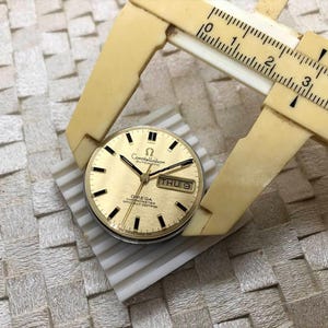 Puede incluir: Una esfera de reloj autom&aacute;tico Omega Constellation dorada con marcadores de hora y agujas negras. La esfera muestra el d&iacute;a y la fecha. Un calibrador beige vintage mide la esfera, que est&aacute; sobre una superficie blanca.