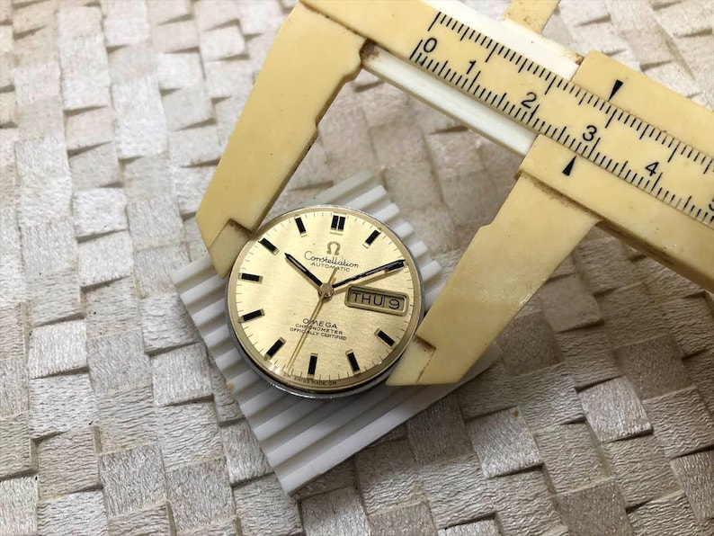 Puede incluir: Una esfera de reloj autom&aacute;tico Omega Constellation dorada con marcadores de hora y agujas negras. La esfera muestra el d&iacute;a y la fecha. Un calibrador beige vintage mide la esfera, que est&aacute; sobre una superficie blanca.