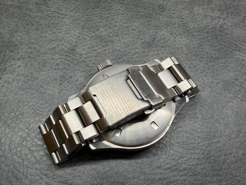May include: A stainless steel wristwatch featuring a brushed metal link bracelet with a fold-over clasp. The watch case back is circular and metallic, showing a winding crown on the side. The design is utilitarian with a polished silver finish.