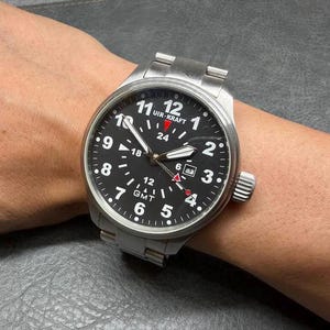 May include: A stainless steel wristwatch with a black dial featuring white Arabic numerals, a date window at the three o'clock position, and a GMT function. The watch face displays the text UIR-KRAFT and includes a red-tipped GMT hand.
