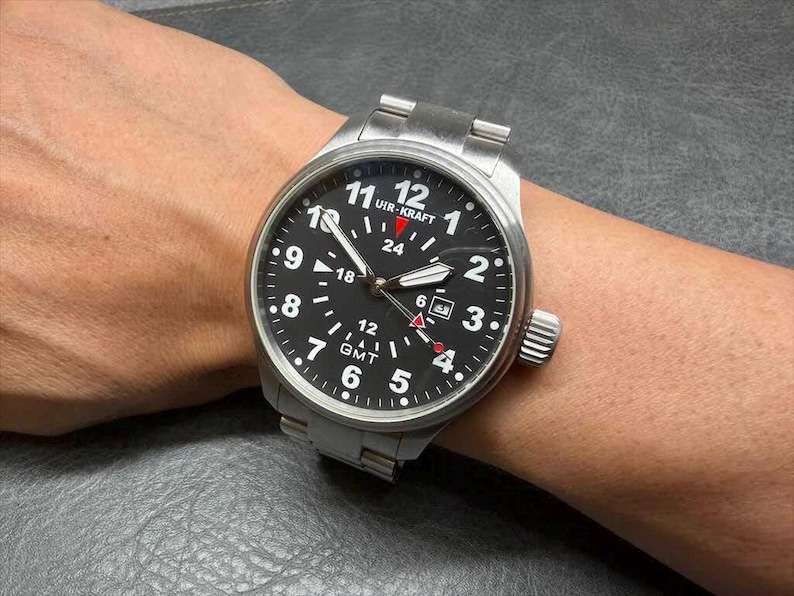 May include: A stainless steel wristwatch with a black dial featuring white Arabic numerals, a date window at the three o'clock position, and a GMT function. The watch face displays the text UIR-KRAFT and includes a red-tipped GMT hand.