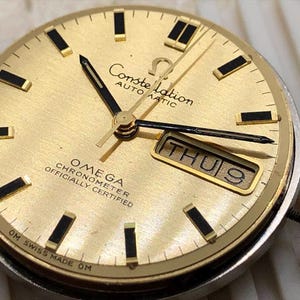 Puede incluir: Una esfera de reloj Omega Constellation dorada con manecillas y marcadores negros. La esfera muestra el d&iacute;a y la fecha, con "THU 9" visible. El reloj est&aacute; marcado con "OMEGA CHRONOMETER OFFICIALLY CERTIFIED" y "OM SWISS MADE OM".