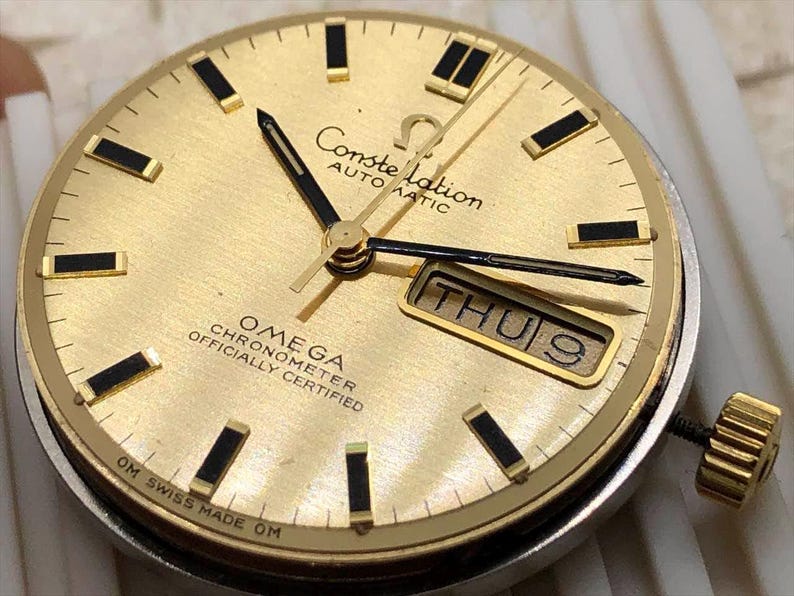 Puede incluir: Una esfera de reloj Omega Constellation dorada con manecillas y marcadores negros. La esfera muestra el d&iacute;a y la fecha, con "THU 9" visible. El reloj est&aacute; marcado con "OMEGA CHRONOMETER OFFICIALLY CERTIFIED" y "OM SWISS MADE OM".