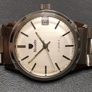 Puede incluir: Un reloj de pulsera Nivada plateado con esfera blanca, marcadores negros y ventana de fecha a las 12. El reloj tiene una correa plateada y corona a las 3. La esfera indica "17 Jewels" y "Swiss Made".
