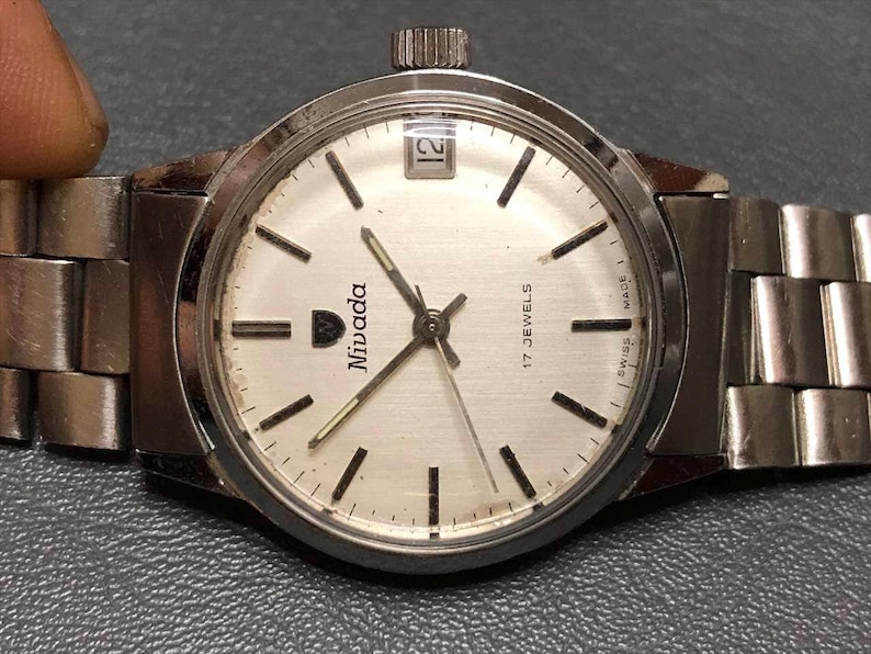 Puede incluir: Un reloj de pulsera Nivada plateado con esfera blanca, marcadores negros y ventana de fecha a las 12. El reloj tiene una correa plateada y corona a las 3. La esfera indica "17 Jewels" y "Swiss Made".