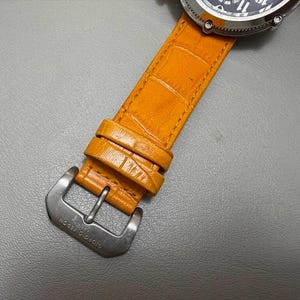 以下が含まれることがあります： シルバーケースと黒い文字盤の腕時計のクローズアップ。時計は、シルバーのバックルが付いた明るいオレンジ色の革ストラップが付いています。ストラップは質感があり、エンボス加工された革で作られているようです。バックルには「GIORGIO FEDON」の文字が刻まれています。