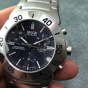 May include: A silver Sector 540 chronograph watch with a dark blue face. The watch features a sapphire crystal, a date display, and is water-resistant to 100 meters (330 ft). The watch has a stainless steel band and bezel with numerals.