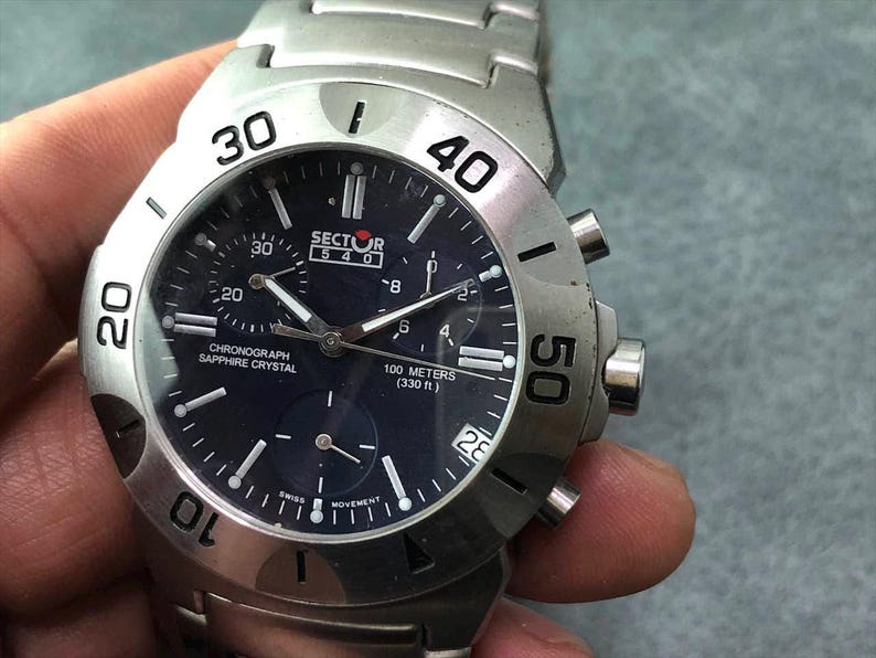 May include: A silver Sector 540 chronograph watch with a dark blue face. The watch features a sapphire crystal, a date display, and is water-resistant to 100 meters (330 ft). The watch has a stainless steel band and bezel with numerals.