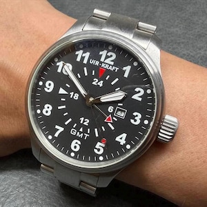 May include: A stainless steel Uir-Kraft GMT wristwatch with a black dial, white Arabic numerals, a date window at the three o'clock position, luminous hands, and a red-tipped GMT hand. The watch features a brushed metal link bracelet.