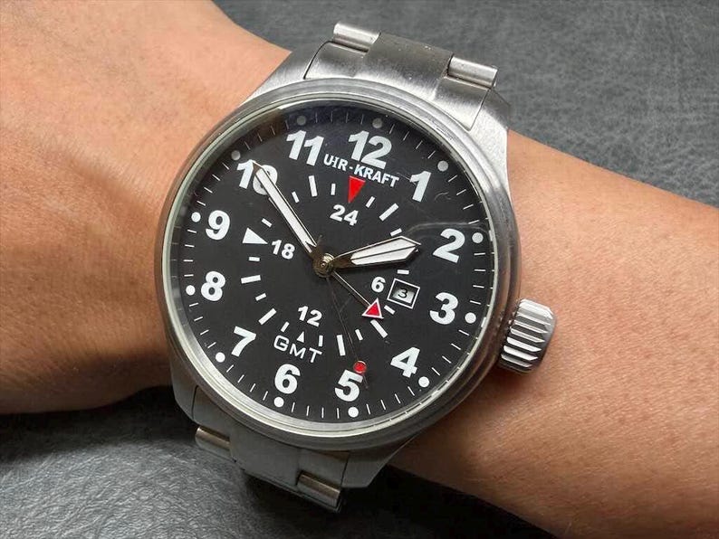 May include: A stainless steel Uir-Kraft GMT wristwatch with a black dial, white Arabic numerals, a date window at the three o'clock position, luminous hands, and a red-tipped GMT hand. The watch features a brushed metal link bracelet.