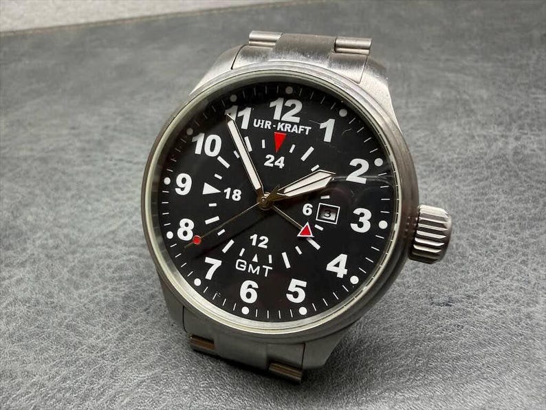 May include: A stainless steel UHR-KRAFT wristwatch with a black dial, white Arabic numerals, and a date window at the three o'clock position. The watch features a GMT function with a red-tipped hand, luminous hands, and a brushed metal link bracelet.
