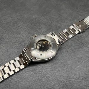 May include: A silver-toned stainless steel wristwatch featuring a round case with an exhibition case back that reveals the internal mechanical movement. The watch includes a matching metal link bracelet with a secure folding deployment clasp.