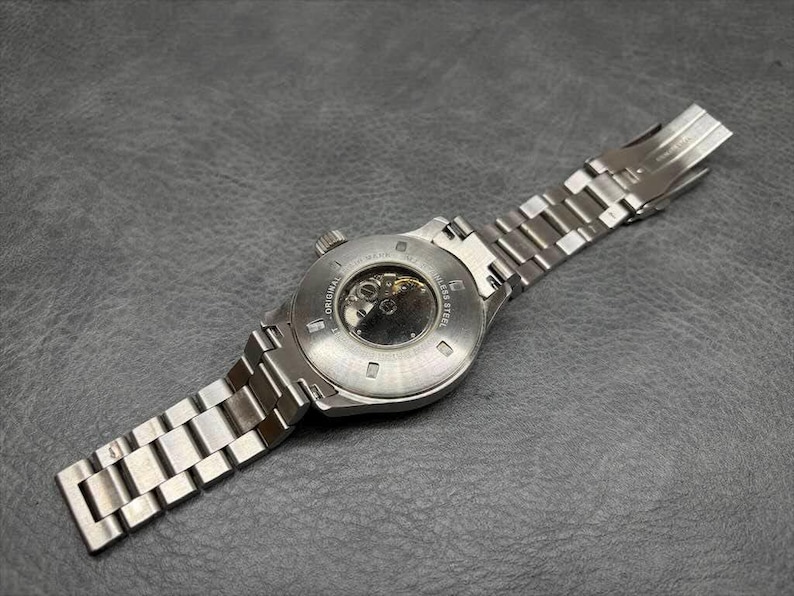 May include: A silver-toned stainless steel wristwatch featuring a round case with an exhibition case back that reveals the internal mechanical movement. The watch includes a matching metal link bracelet with a secure folding deployment clasp.