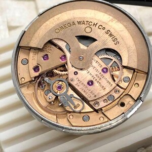 Puede incluir: Primer plano del movimiento de un reloj Omega. El mecanismo del reloj presenta engranajes dorados, rub&iacute;es y el texto "OMEGA WATCH CO SWISS". El movimiento est&aacute; etiquetado como "TWENTY-FOUR 24 JEWELS".