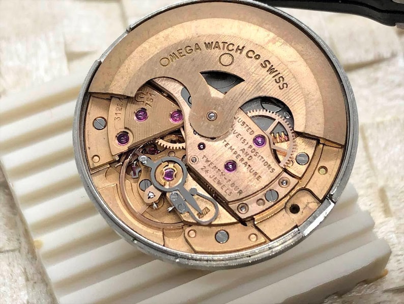 Puede incluir: Primer plano del movimiento de un reloj Omega. El mecanismo del reloj presenta engranajes dorados, rub&iacute;es y el texto "OMEGA WATCH CO SWISS". El movimiento est&aacute; etiquetado como "TWENTY-FOUR 24 JEWELS".