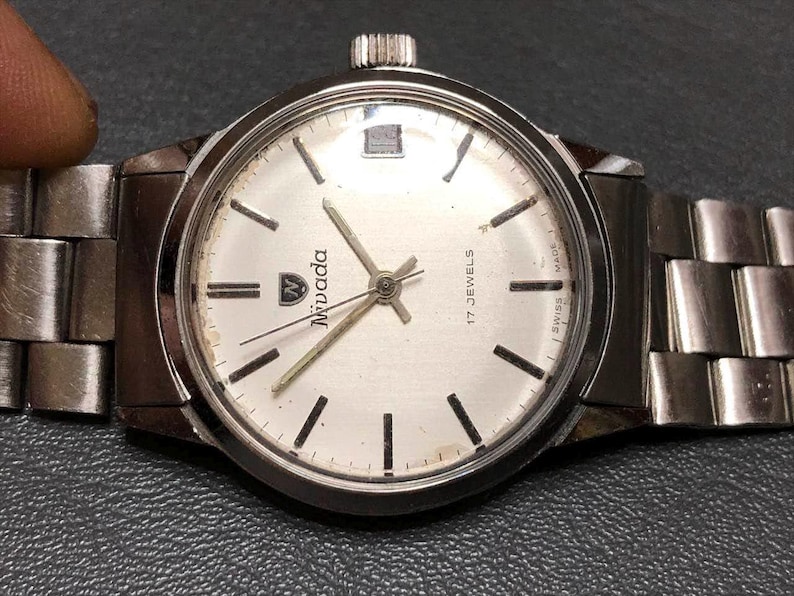 Puede incluir: Reloj de pulsera Nivada plateado con esfera blanca, marcadores de hora negros y agujas plateadas. El reloj tiene una correa de acero inoxidable y una corona a las 3 en punto. La esfera del reloj dice "Nivada" y "17 Jewels Swiss Made".