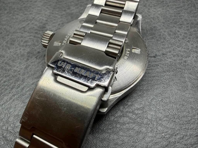 May include: A silver-toned stainless steel wristwatch featuring a metal link bracelet and a fold-over clasp engraved with the text UIR-KRAFT. The circular case back displays the text 5ATM, indicating water resistance, along with other markings.