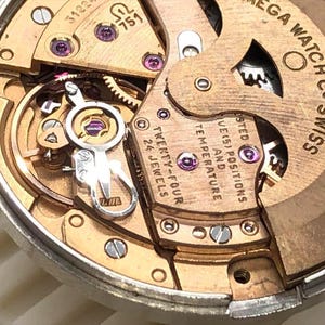 Puede incluir: Primer plano de un movimiento de reloj con engranajes, tornillos y joyas doradas. El mecanismo del reloj tiene el texto "OMEGA WATCH CO SWISS" y "TWENTY-FOUR 24 JEWELS". Un dispositivo mec&aacute;nico complejo.