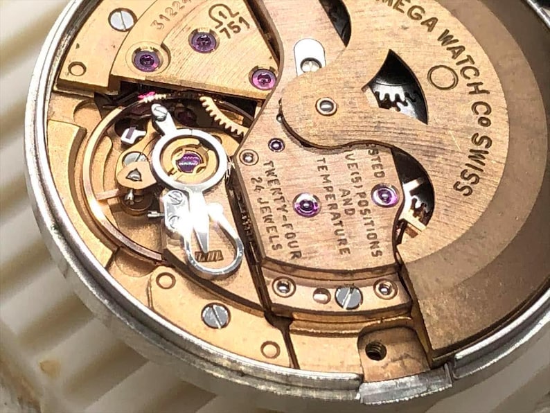 Puede incluir: Primer plano de un movimiento de reloj con engranajes, tornillos y joyas doradas. El mecanismo del reloj tiene el texto "OMEGA WATCH CO SWISS" y "TWENTY-FOUR 24 JEWELS". Un dispositivo mec&aacute;nico complejo.