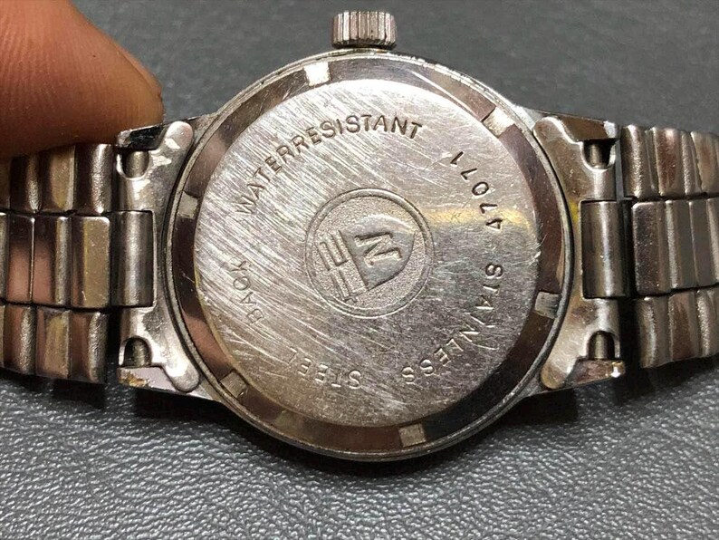 Puede incluir: Primer plano de la parte trasera de un reloj de pulsera plateado con una correa de acero inoxidable. La esfera del reloj es circular con las palabras "WATER RESISTANT" y "STAINLESS STEEL" grabadas. El reloj tambi&eacute;n tiene el n&uacute;mero "47011" grabado.