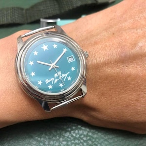 May include: A silver wristwatch with a teal face featuring white stars and the text "Nancy M. Lee". The watch has silver hands and a date display. The watch is on a wrist.