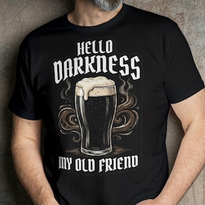 May include: Black t-shirt with the text "Hello Darkness My Old Friend" and a graphic of a beer glass with foam. The shirt is worn by a person in blue jeans. The background is a textured, neutral-toned wall.