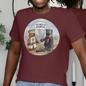 May include: Burgundy t-shirt with a cartoon of two knights, one in white and one in black armour, with swords. A speech bubble says "'Tis but a scratch". The person is wearing blue jeans.