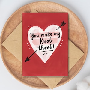 Puede incluir: Tarjeta de felicitación roja con un corazón blanco y el texto "You make my Knob throb!" atravesada por una flecha negra. La tarjeta descansa sobre una bandeja de madera con un sobre marrón.