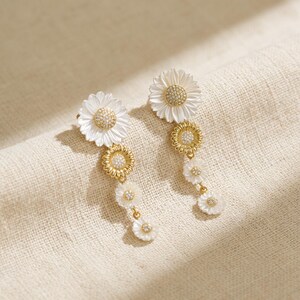 May include: A pair of dangling earrings featuring a daisy design. Each earring has a large white flower at the top, followed by a gold-colored flower and smaller white flowers. The center of each flower is embellished with small, sparkling stones.