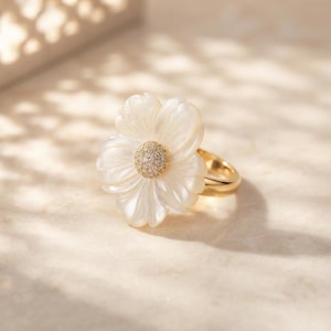 May include: A gold ring featuring a large, white, carved mother-of-pearl flower as the centerpiece. The center of the flower is adorned with a cluster of small, sparkling clear stones. The band of the ring is made of polished gold metal.