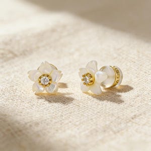 Könnte beinhalten: Ein Paar blütenförmige Ohrstecker. Jeder Ohrring hat ein weißes, blütenblattartiges Design mit einem goldfarbenen Mittelteil und einem kleinen, klaren Edelstein. Die Ohrringe sind vor einem neutralen, strukturierten Hintergrund platziert.