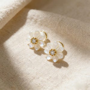 Op de afbeelding: Een paar bloemvormige oorbellen. Elke oorbel heeft een wit, bloembladachtig ontwerp rond een centrale, heldere edelsteen. De oorbellen hebben goudkleurige accenten en zijn geplaatst tegen een neutrale, getextureerde achtergrond.