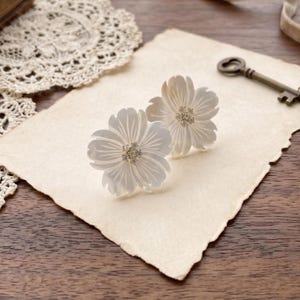 May include: Two flower-shaped earrings crafted from iridescent white shell material. Each earring features five carved petals with delicate linear detailing and a central cluster of small, clear, sparkling stones set in a metallic base.