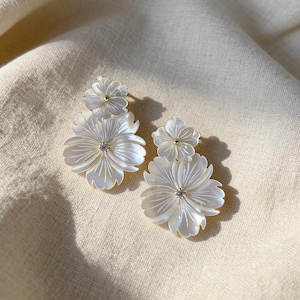 Puede incluir: Un par de pendientes blancos con forma de flor. Cada pendiente presenta una flor grande de múltiples pétalos con una pequeña gema brillante en el centro. Una flor más pequeña se encuentra encima de la más grande. Los pendientes tienen un poste dorado.