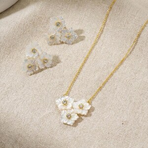 Può includere: Un set di collana e orecchini abbinati color oro. La collana presenta tre ciondoli a forma di fiore bianco con un piccolo cristallo al centro. Gli orecchini hanno tre ciondoli a forma di fiore bianco. I gioielli sono su un tessuto beige.
