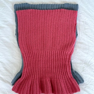 May include: A handmade, sleeveless crochet top in a vibrant red hue, with a contrasting gray trim. The top features a ribbed texture and a flared peplum hem, showcasing a unique, handcrafted design. The garment is laid flat on a white surface.