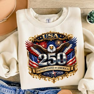 May include: Cream-colored sweatshirt featuring a patriotic design. The graphic includes a majestic eagle with outstretched wings, the number "250", American flags, and the text "USA" and "United States of America 1776-2026".