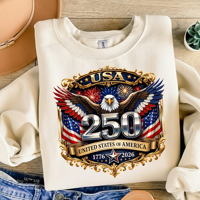 May include: Cream-colored sweatshirt featuring a patriotic design. The graphic includes a majestic eagle with outstretched wings, the number "250", American flags, and the text "USA" and "United States of America 1776-2026".