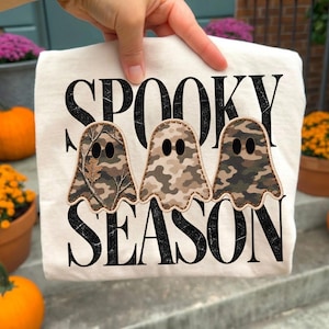 Könnte beinhalten: Ein cremefarbenes T-Shirt mit dem schwarzen, abgenutzten Schriftzug SPOOKY SEASON. Drei geisterförmige Aufnäher sind mittig appliziert, jeder mit einem anderen Tarnmuster in Braun-, Grün- und Beigetönen sowie schwarzen Augen.