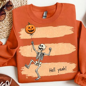May include: An orange crewneck sweatshirt featuring a graphic of a white skeleton holding a string attached to an orange jack-o'-lantern balloon. The design includes three horizontal light orange brushstroke stripes and the text Hell yeah!