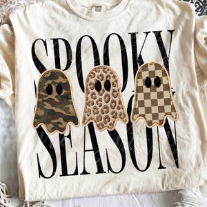 May include: A cream-coloured t-shirt featuring the black text SPOOKY SEASON. Three ghost-shaped graphics are centred on the shirt, each with black oval eyes. The ghosts feature camouflage, leopard print, and a brown and cream checkerboard pattern.