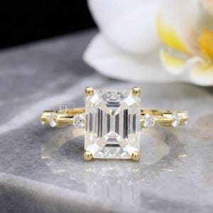 Könnte beinhalten: Ein goldener Ring mit einem klar geschliffenen Smaragd als Mittelstück. Das Band hat kleinere, runde, klare Edelsteine. Der Ring ist vor einem verschwommenen Hintergrund mit einer weißen Blume und einer grauen Oberfläche platziert.