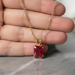 Puede incluir: Un collar dorado con un colgante rectangular de piedra preciosa rojo intenso. El colgante está engastado en un bisel dorado y cuelga de una delicada cadena dorada. El collar se muestra sobre un fondo neutro.