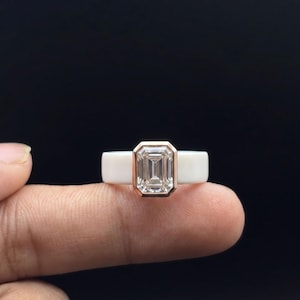 Peut inclure: Une bague composée d'un anneau en céramique blanche et d'une pierre transparente taille émeraude, sertie dans une monture en or rose. La pierre rectangulaire est facettée et reflète la lumière, contrastant avec l'anneau blanc lisse.