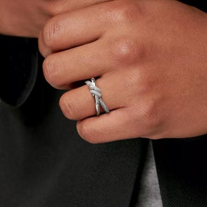 K&ouml;nnte beinhalten: Ein silberfarbener Ring mit Knoten-Design. Der Ring hat kleine, funkelnde Steine entlang eines Bandes und am Knoten. Der Ring wird an einem Finger getragen, mit dunklem Hintergrund.