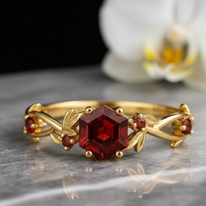 Könnte beinhalten: Ein goldener Ring mit einem großen, sechseckigen, tiefroten Edelstein im Zentrum. Das Band ist mit filigranen Blatt- und Rankendetails sowie kleineren roten Edelsteinen verziert. Der Ring ist vor einem unscharfen Hintergrund platziert.