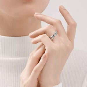 K&ouml;nnte beinhalten: Ein silberfarbener Ring mit einem Knoten-Design, verziert mit kleinen, funkelnden Steinen. Der Ring befindet sich am Finger einer Person und zeigt seine komplizierten Details und seinen eleganten Stil. Das Design des Rings ist eine moderne Interpretation eines klassischen Knotens.