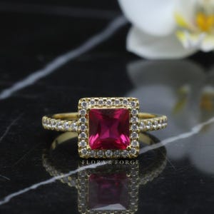 May include: A gold ring featuring a square, ruby-red central gemstone, encircled by a halo of small white stones. The band is also embellished with tiny white stones. The ring is set against a black and white marble surface. The text "FLORA & FORGE" is visible.